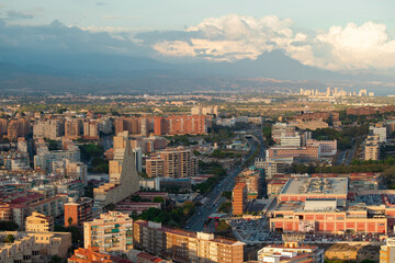 A panoramic aerial view of a bustling city with modern buildings, a busy road, and a scenic mountain range in the background under a partly cloudy sky. Alicante, Spain
