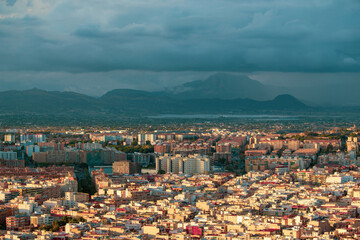 A panoramic view of a densely populated city with a mountain range in the background, under a cloudy sky. Captures the blend of urban development and natural beauty. Alicante, Spain.