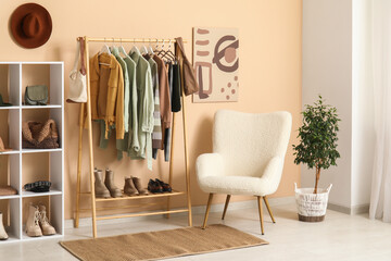 Rack with clothes, armchair and shelving unit near beige wall in dressing room
