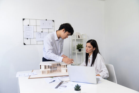 Professional male asian architect using ruler to measure house model length while young beautiful caucasian colleague using laptop to analyzed data on meeting table with house model. Immaculate. - Powered by Adobe