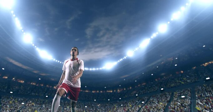 Football player, wearing red and white uniform, kicks the ball in a jump, in slow motion on the professional football stadium with animated crowd.