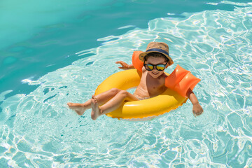 Young boy enjoying a sunny day in the pool, wearing sunglasses, a hat, and arm floats while relaxing on a yellow inflatable ring. Space for text