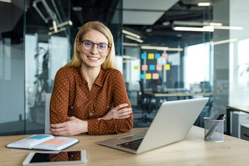 Mature business woman sitting confidently in modern office, with laptop and tablet on desk. She exudes professionalism, leadership, success, ideal for corporate or office themes.