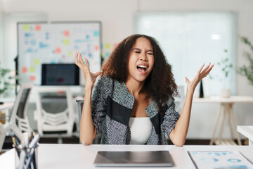 Overwhelmed businesswoman yelling with raised hands at her desk, expressing frustration and stress...