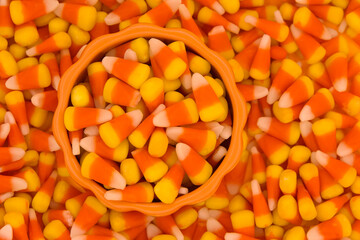 Top view of a small porcelain pumpkin bowl filled with candy corn spilling over onto a background of the yellow orange and white striped traditional Halloween candy.