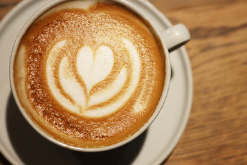 Creamy cappuccino with beautiful flower heart pattern in a white ceramic cup on a wooden table in a coffee shop. Closeup. Copy space. Top view