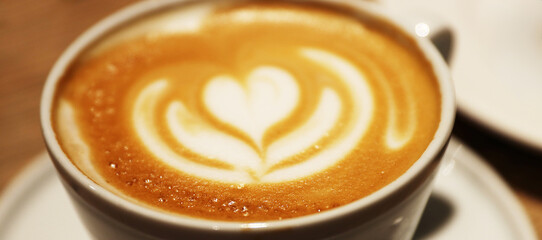 Closeup of creamy cappuccino with beautiful flower heart pattern in a white ceramic cup on a wooden table in a coffee shop. Copy space. Selective focus with bokeh