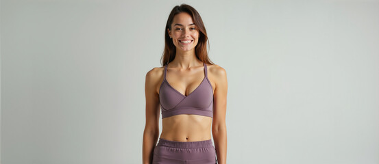 A young woman with long brown hair is smiling at the camera. She is wearing a purple sports bra and purple leggings. She is standing against a white wall. She has a toned physique and is looking confi