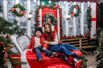 Two siblings boy and girl sit on a vintage red car in front of a festive house decorated with wreaths, garlands, and Christmas lights. The cozy winter scene exudes holiday cheer.