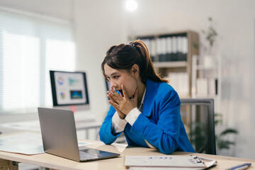 Young businesswoman with a surprised expression is sitting at her desk, covering her mouth with her hands while looking at her laptop, reacting to unexpected news or data