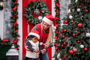 Caucasian mother and little African American daughter, both in Santa hats, enjoy bonding while decorating a festive Christmas tree outdoors, surrounded by holiday decor and a vintage red truck.