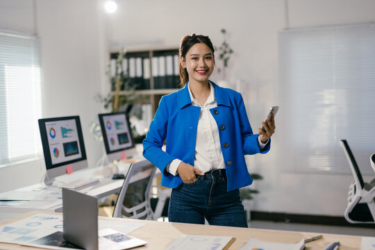 Asian businesswoman is holding a smartphone and smiling while standing in a modern office with computers and documents on desks