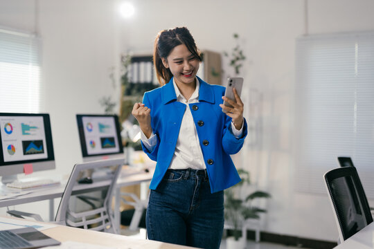 Asian businesswoman is expressing excitement while looking at her smartphone in a modern office, celebrating a positive outcome or achievement