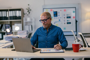 Confident mature entrepreneur sitting at desk in modern office, using laptop and examining financial reports, working with concentration and focus on project