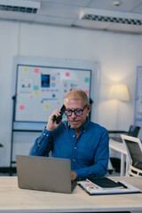 Focused mature businessman sitting at desk in modern office, using laptop and having phone conversation, demonstrating efficiency and multitasking skills in corporate environment