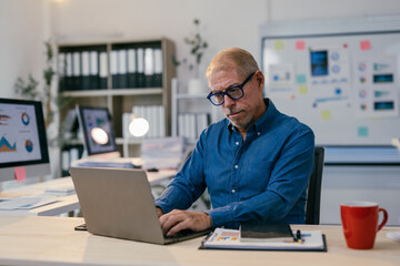 Senior businessman working on his laptop in a modern office, analyzing charts and data displayed on computer screens, focused on his tasks
