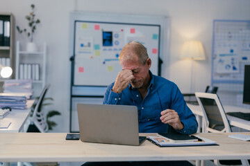 Exhausted mature manager rubbing his nose bridge, suffering from headache or eye strain after long computer work at desk in modern office