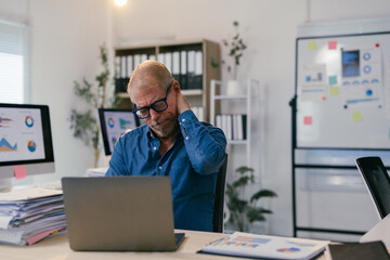 Tired mature entrepreneur experiencing neck pain while sitting at desk in modern office, surrounded...
