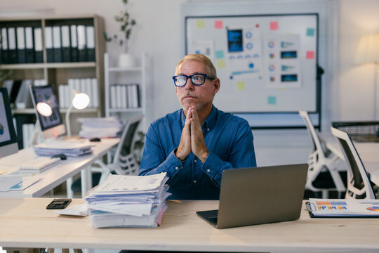 Senior manager sitting at his desk with hands clasped, contemplating a challenging situation amidst a pile of documents and a laptop, in a modern office environment