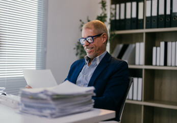 Senior businessman happily working in a modern office, surrounded by documents and a laptop, showcasing professionalism and expertise