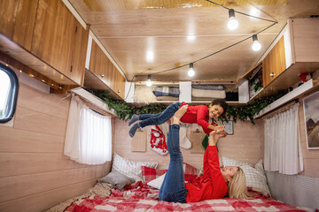 Caucasian Mother and African American son share a joyful moment, playing airplane on a cozy bed in a decorated camper, creating holiday memories together. Smiling woman lifting her child