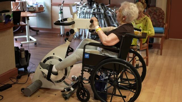 An elderly woman with white hair sits in a wheelchair and uses a stationary bike for exercise. She has a bandage on her arm and a yellow bracelet. 