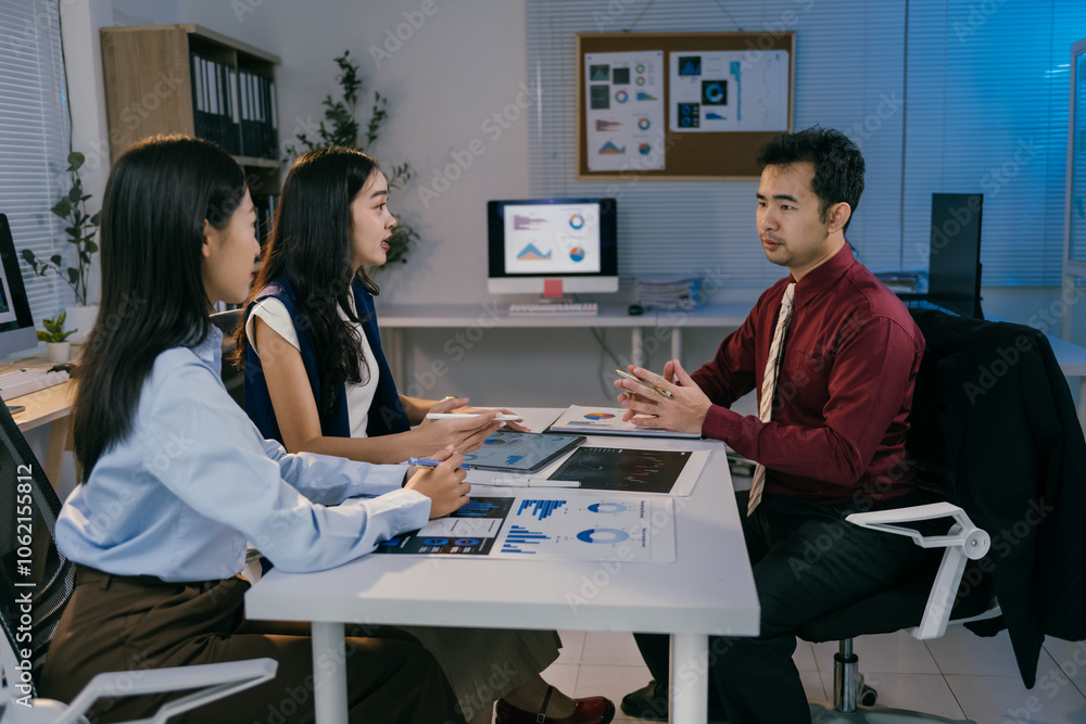 Poster three asian businesspeople sitting at desk in modern office at night, discussing charts and data dis