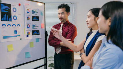 Asian businesspeople discussing marketing strategy during a presentation with colorful pie charts and bar graphs displayed on a whiteboard, fostering collaboration and data-driven decision-making