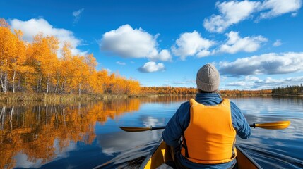 A person in a yellow kayak paddles gently across a tranquil lake, surrounded by stunning autumn colors and a vibrant blue sky dotted with clouds. The scenery is picturesque and peaceful