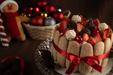 Festive Christmas Charlotte cake adorned with strawberries, cherries, chocolate truffles, and whipped cream, surrounded by ladyfingers and a red ribbon, evoking holiday cheer and winter celebrations.