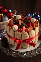 Festive Christmas Charlotte cake adorned with strawberries, cherries, chocolate truffles, and whipped cream, surrounded by ladyfingers and a red ribbon, evoking holiday cheer and winter celebrations.