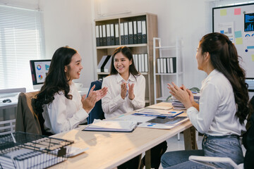 Three young businesswomen are sitting at a desk in a modern office, clapping their hands together after a successful presentation, celebrating their achievements and teamwork