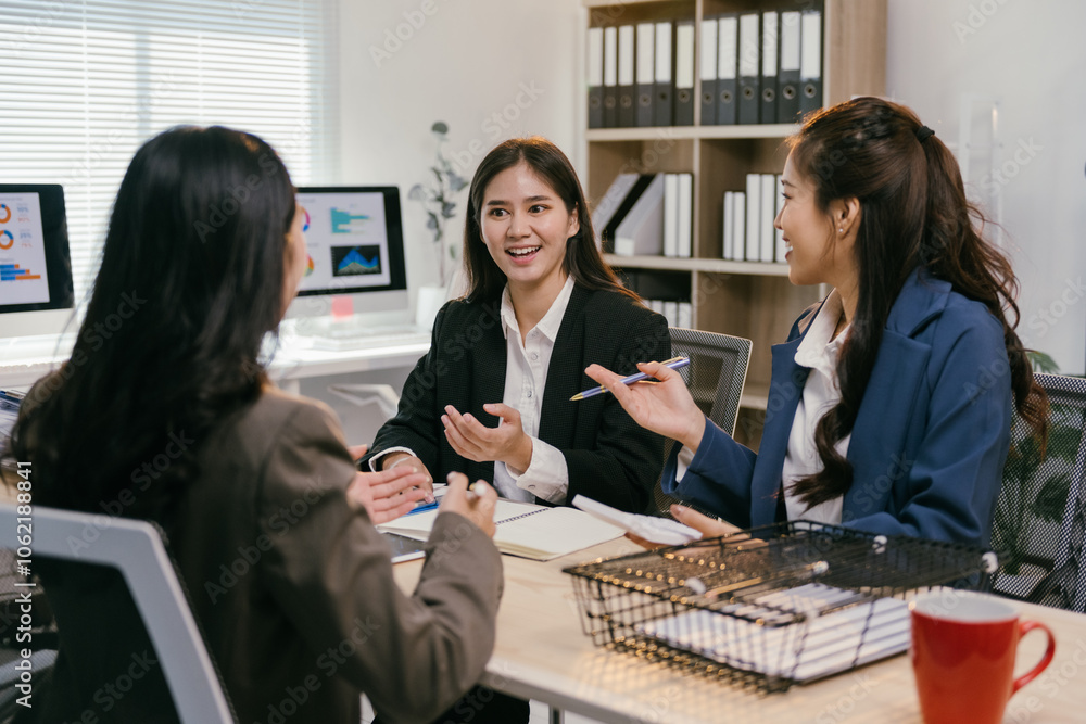Poster three asian businesswomen are sitting at a desk in a modern office, discussing about charts displaye