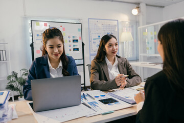 Three asian businesswomen are sitting at a desk in a modern office, collaborating on a project using a laptop and discussing charts and graphs during a productive meeting