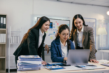Three asian businesswomen are smiling and collaborating on a project, using a laptop and analyzing charts and graphs in a modern office environment