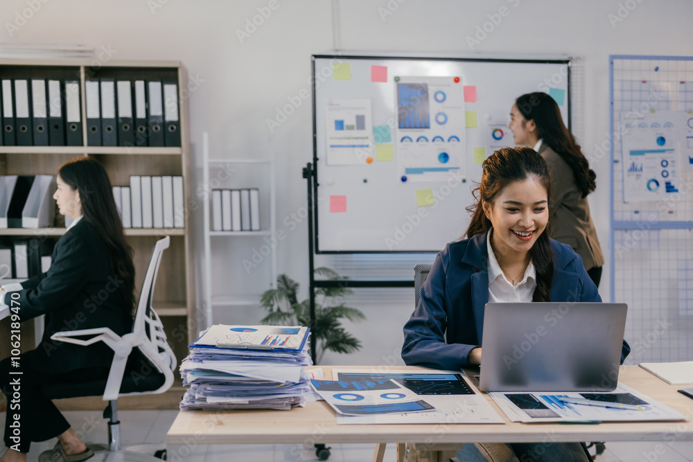 Poster smiling businesswoman working on laptop with charts and graphs in a modern office, her colleagues ar