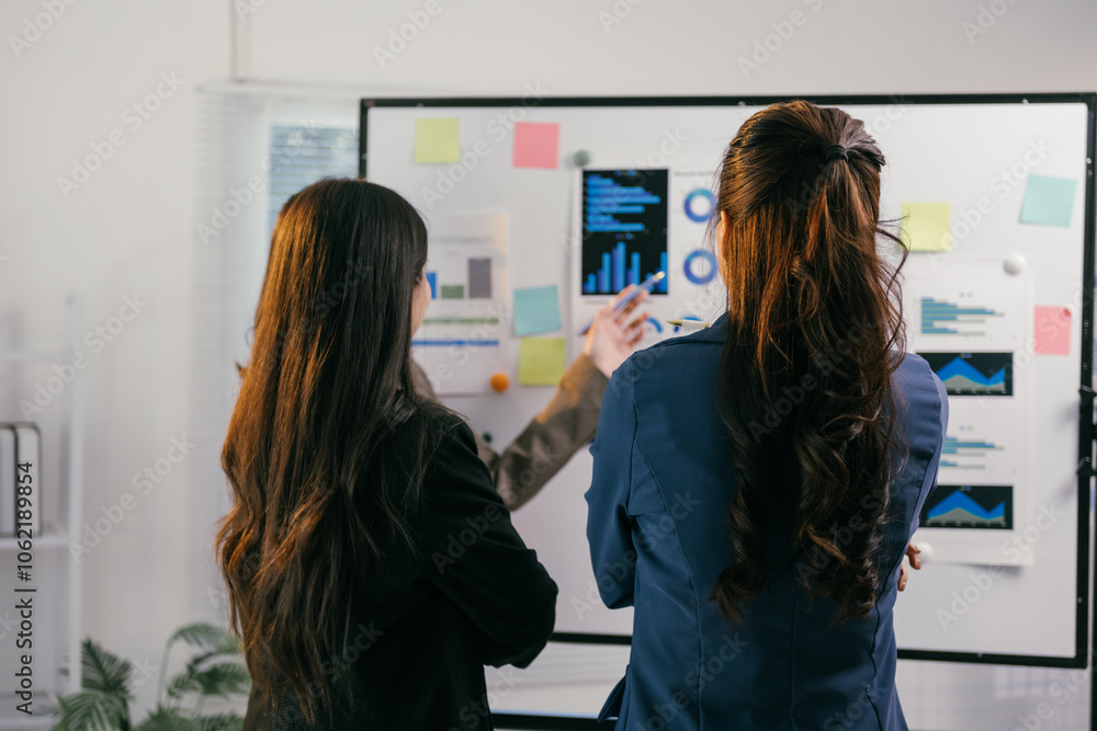 Poster two businesswomen are discussing and analyzing charts and reports displayed on a whiteboard during a