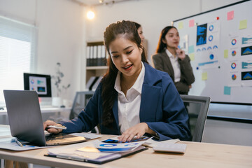 Young businesswoman analyzing financial reports and using laptop, with colleagues discussing charts in background, showcasing teamwork and data analysis in modern office environment
