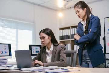 Two young asian businesswomen are working on a laptop computer with a pile of paperwork and having a serious discussion in a modern office