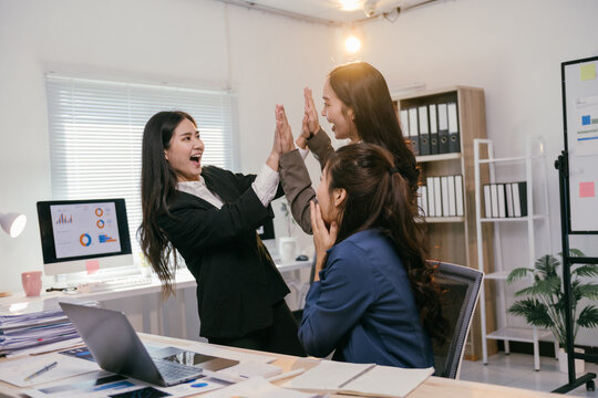 Three excited businesswomen are giving a high five, celebrating their success in the office, surrounded by computers, documents, and office supplies, demonstrating teamwork and achievement
