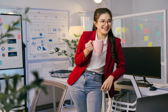 Young manager drinking a coffee mug and smiling while leaning on her chair in her office, with charts and graphs visible on the wall behind her