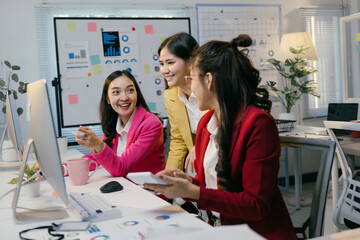 Three asian businesswomen are working together on a computer, discussing and analyzing financial data with smiles and enthusiasm in a modern office environment
