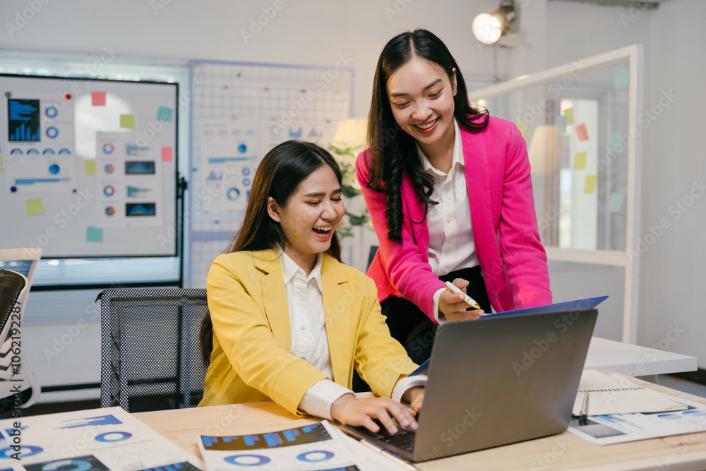 Poster two cheerful asian businesswomen are working together in a modern office, analyzing data on a laptop
