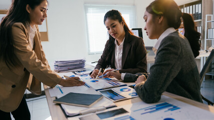 Three asian businesswomen are discussing company performance and analyzing financial charts during a productive meeting in a modern office