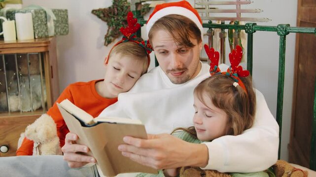 A father in a Christmas hat reads a bedtime story to his children, who wear festive reindeer antlers, creating a heartwarming holiday scene before they fall sleep