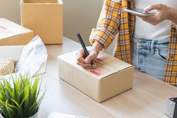 Online seller store, owner small business entrepreneur asian young woman packing product in box, using smartphone, mobile check purchase order from web prepare parcel sent delivery company to customer