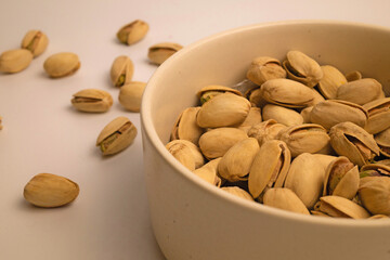 Delicious salted pistachios fill the beige ceramic bowl, some of them spilling out onto the soft beige background.