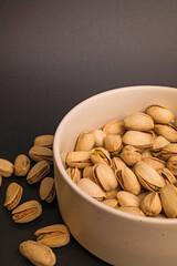 Pistachios lie in a deep white ceramic bowl on a black background, with some scattered around.