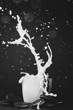 Milk splashing out of a glass cup surrounded by a stack of chocolate cookies