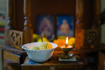 A home altar with a traditional oil lamp, ladoo sweets in a bowl, and blurred deities in the background, symbolizing devotion and spirituality in Hindu culture. Ideal for religious and culture. 